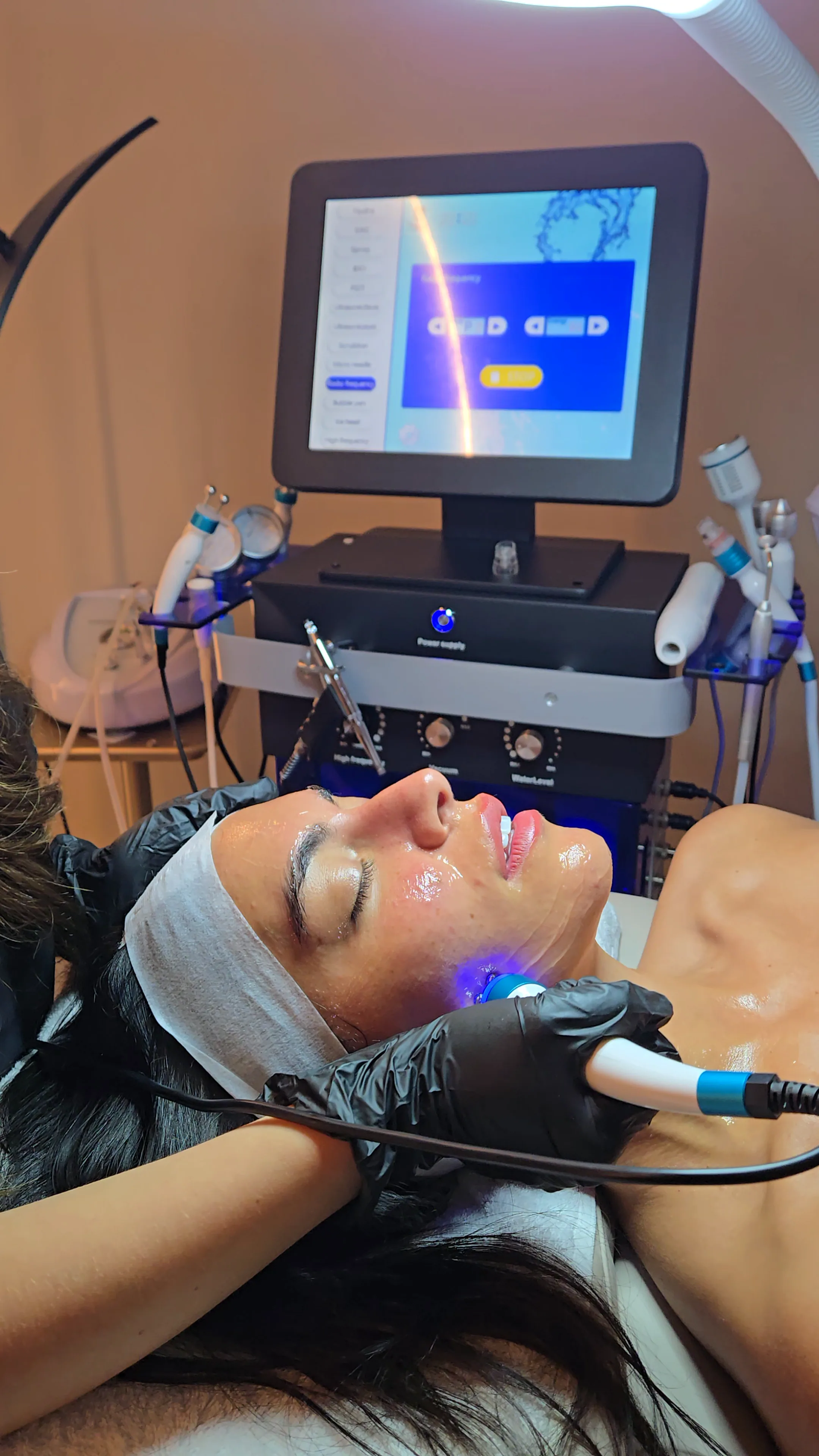 A practitioner holds a blue LED wand to a client's jaw, the facial machine and its handpieces behind her