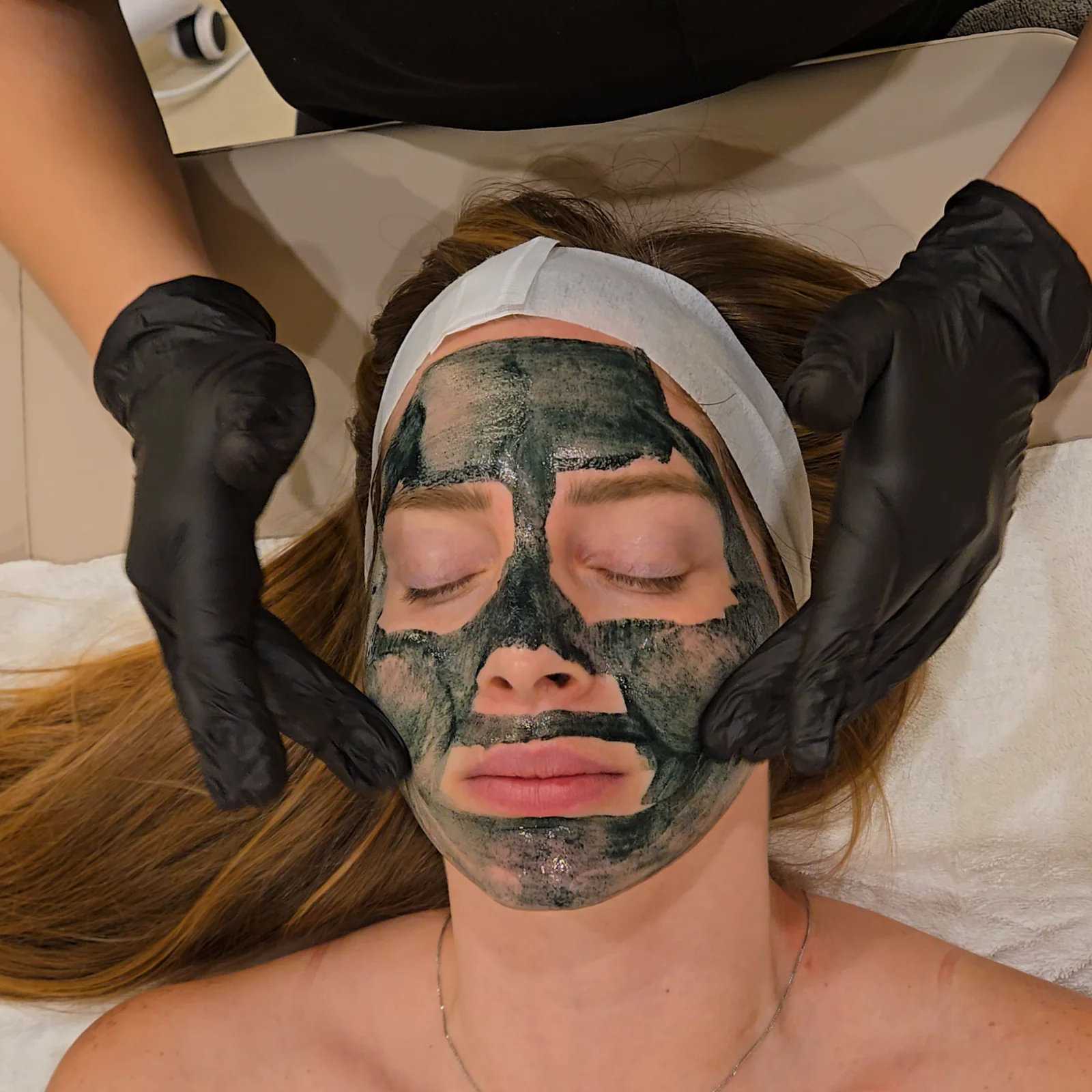 A client with a green herbal peel mask on her face, framed by the practitioner's gloved hands
