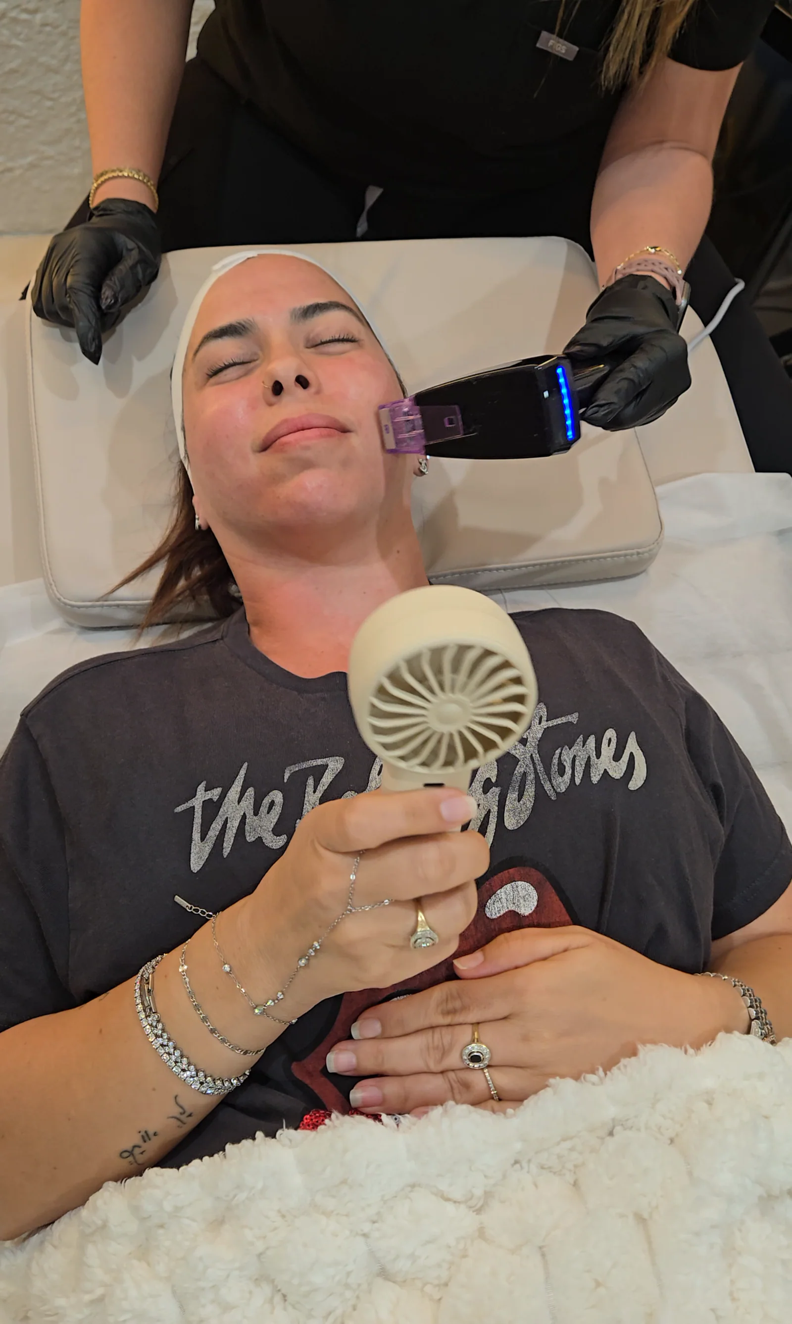 A client holds a small fan and rests her eyes as a practitioner moves an RF microneedling handpiece along her cheek