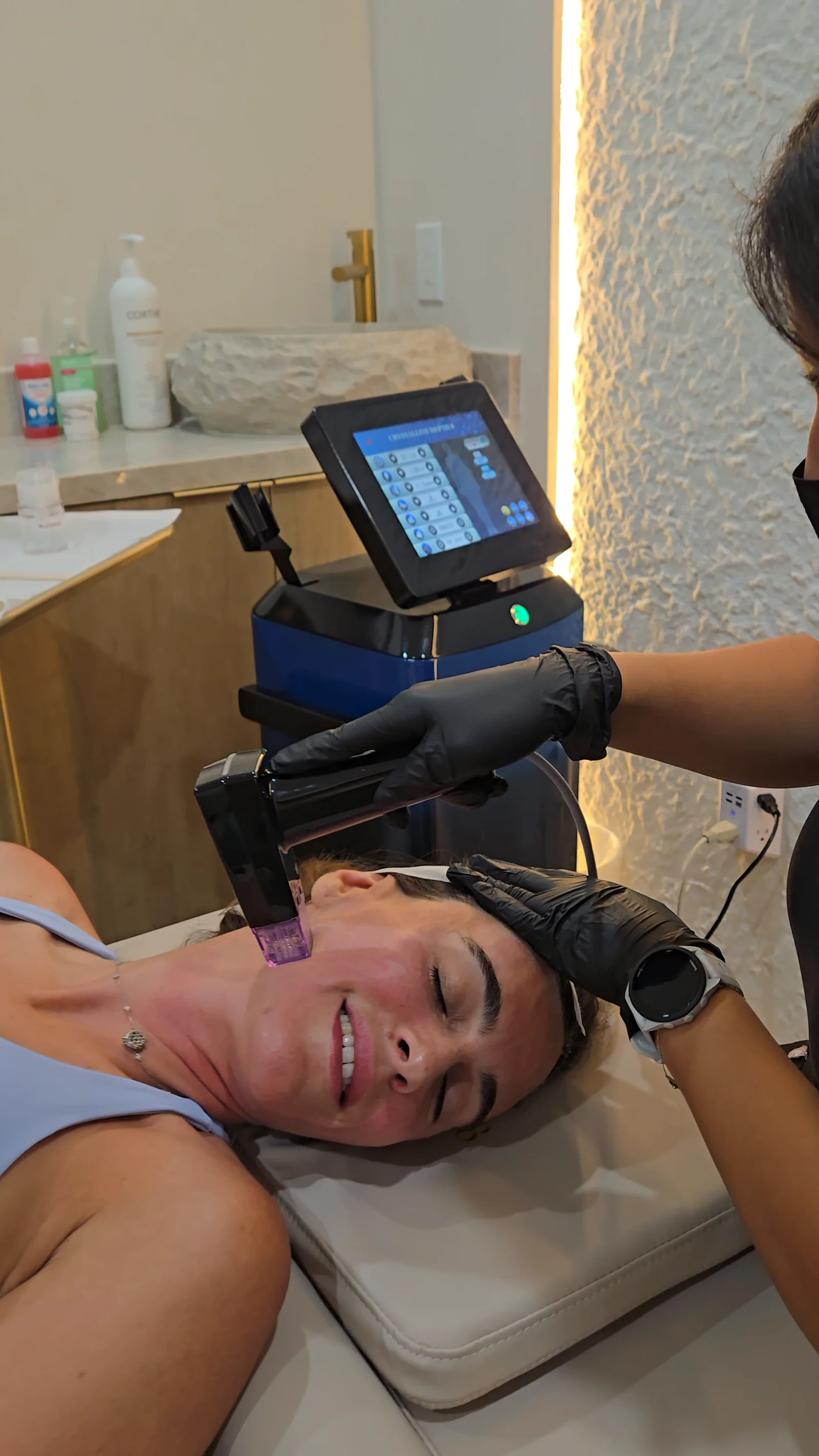 A smiling client during an RF microneedling session, the handpiece on her cheek and the device screen behind her