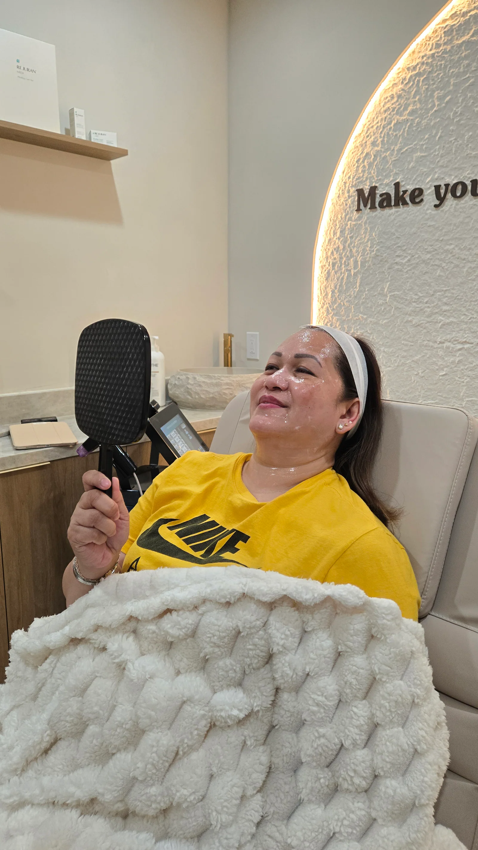 A client under a knit blanket checks her skin in a hand mirror while wearing a sheet mask