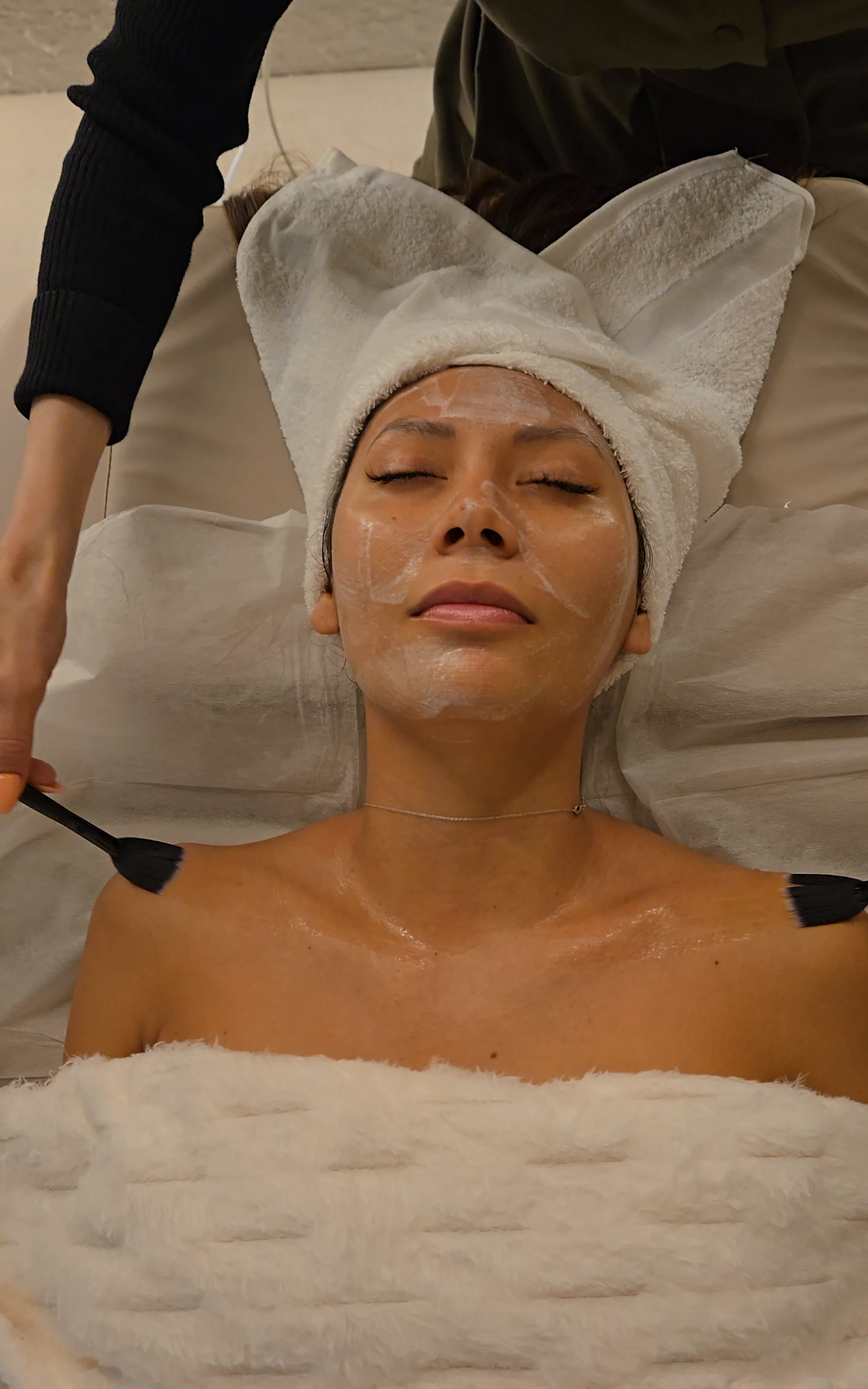 A client rests in a towel turban under a clear facial mask as a practitioner works with two brushes at her shoulders