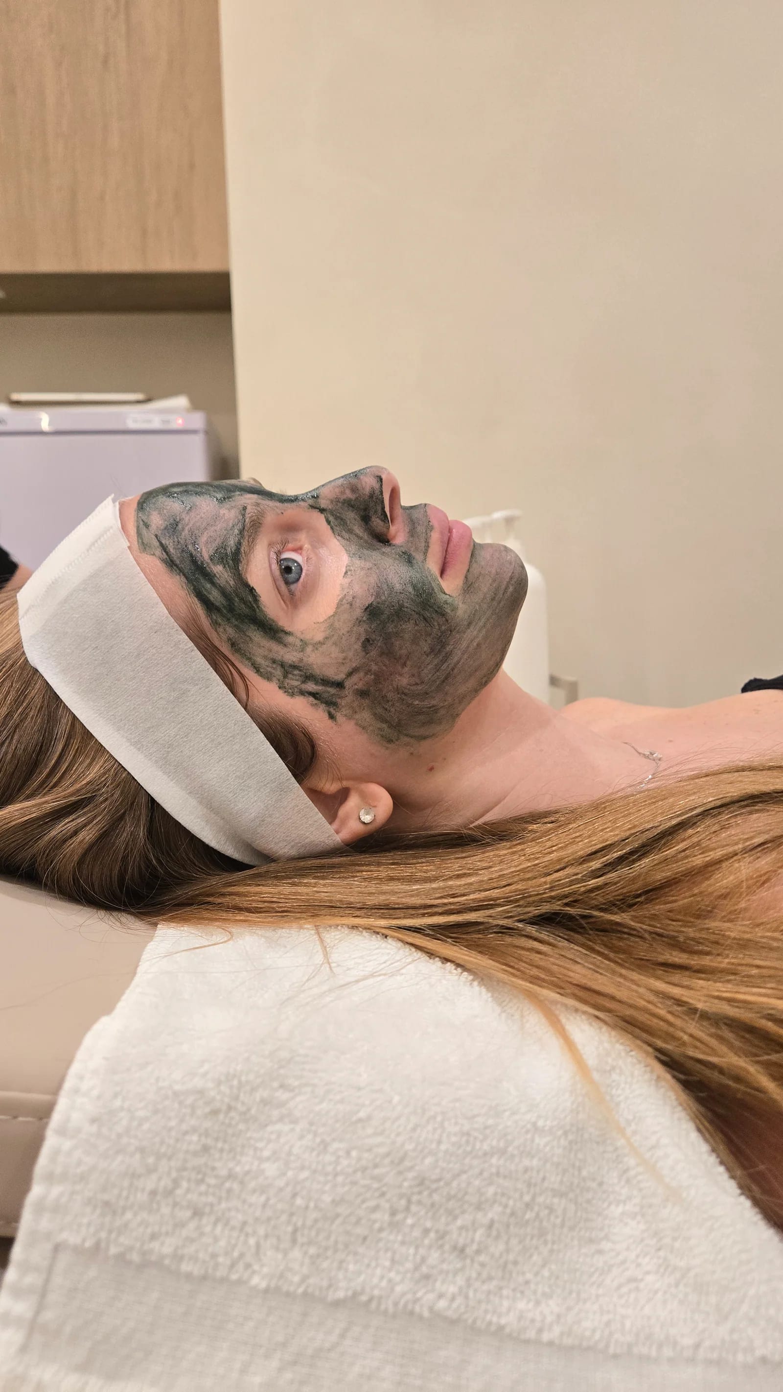 A client rests with a dark green Green Sea Peel mask on her face and a headband in her hair