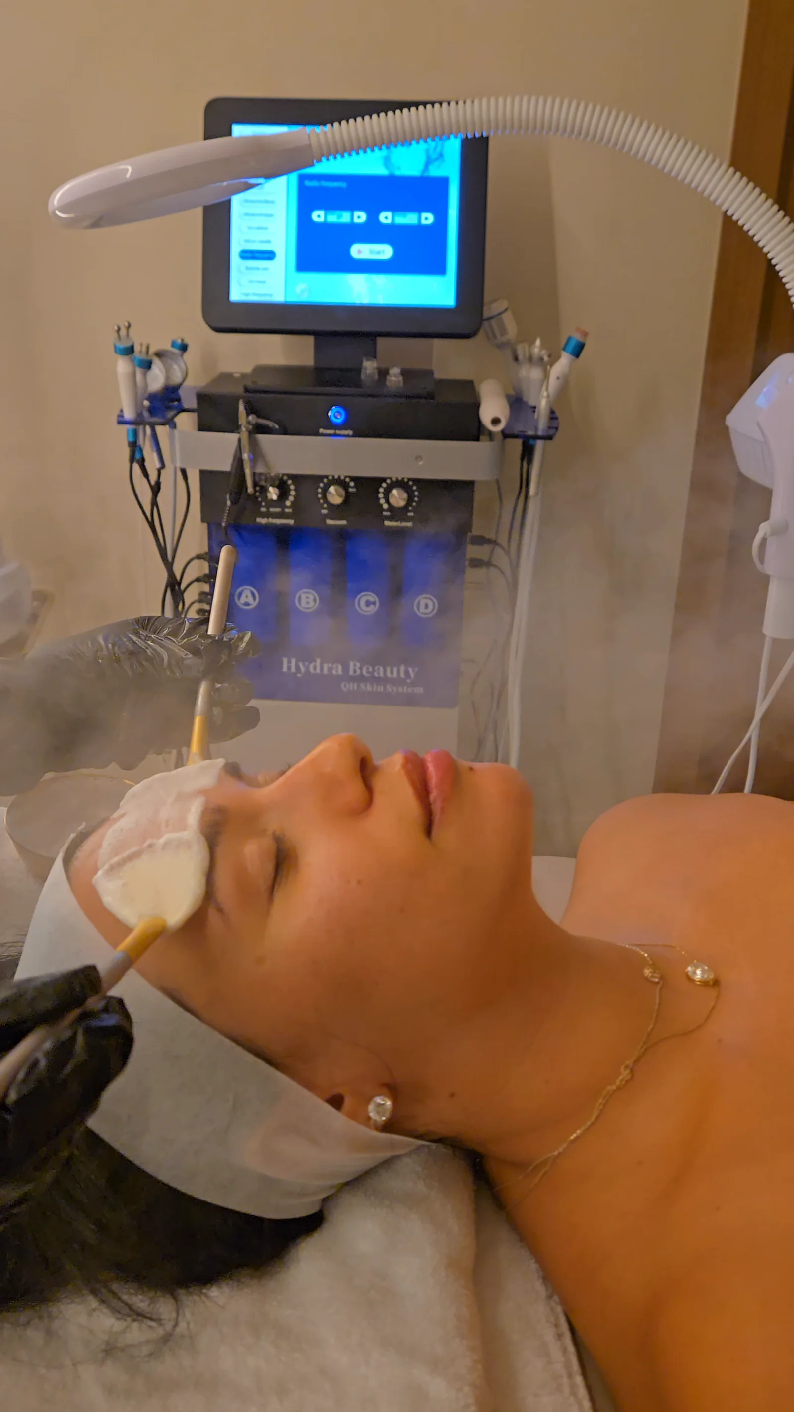 A client resting under a warm mist as a mask is brushed on, the Hydra Beauty facial machine behind her