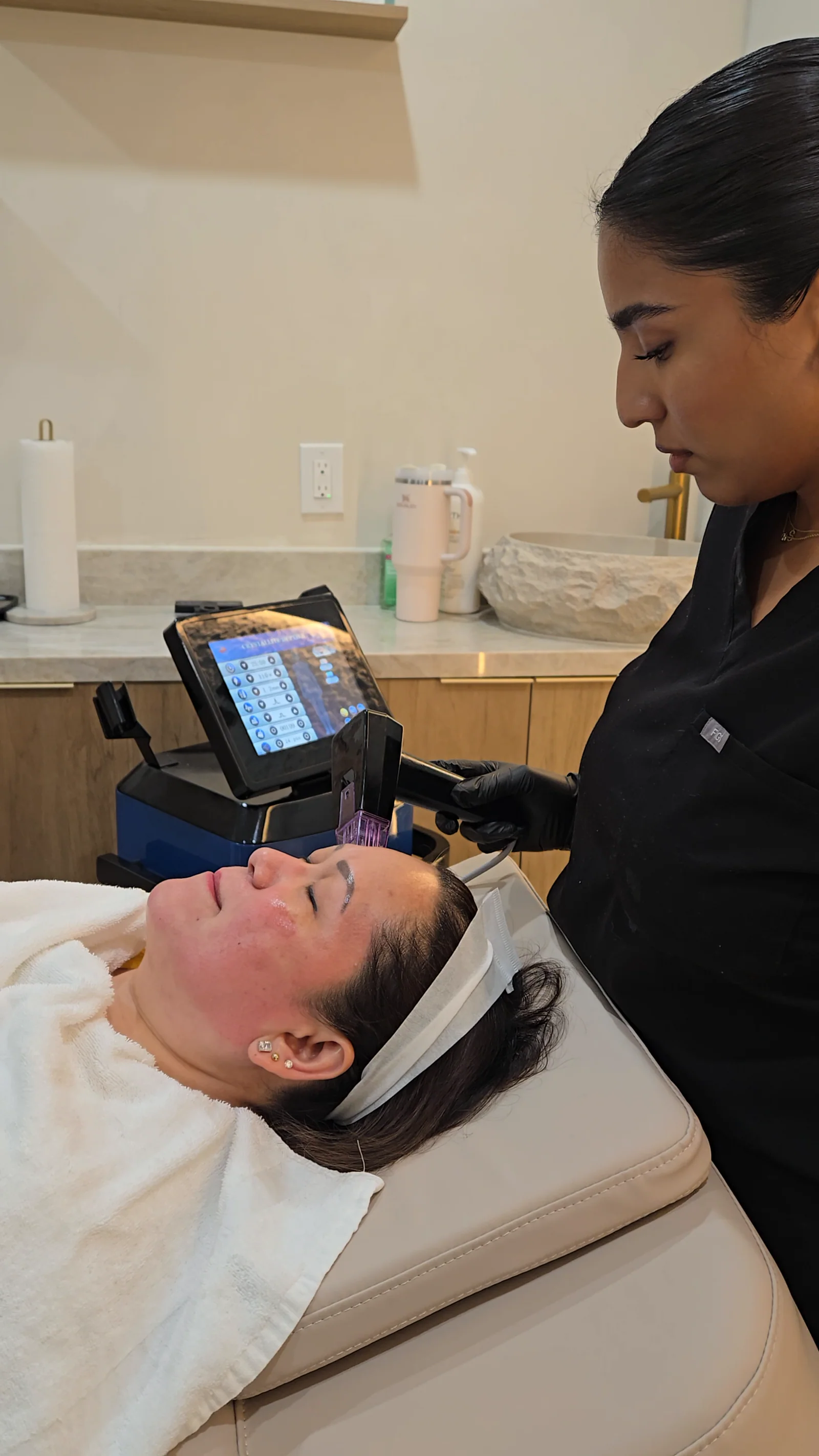 A practitioner holds an RF microneedling handpiece to a client's forehead, the device screen beside the bed